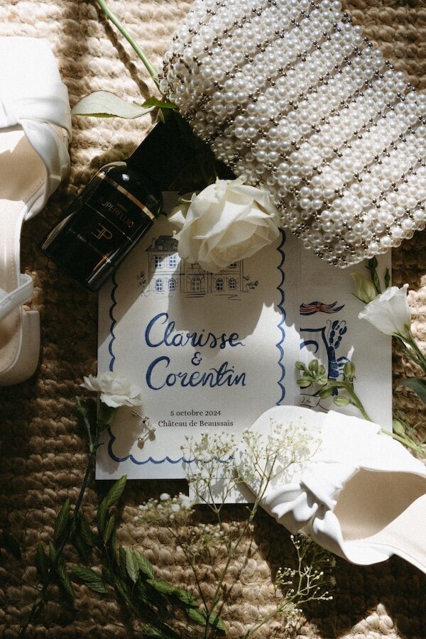 Accessoires de la mariée disposés avec soin : carton d'invitation illustré, sac perlé, parfum et fleurs. Par Kamescop, photographe mariage vintage en Bretagne.