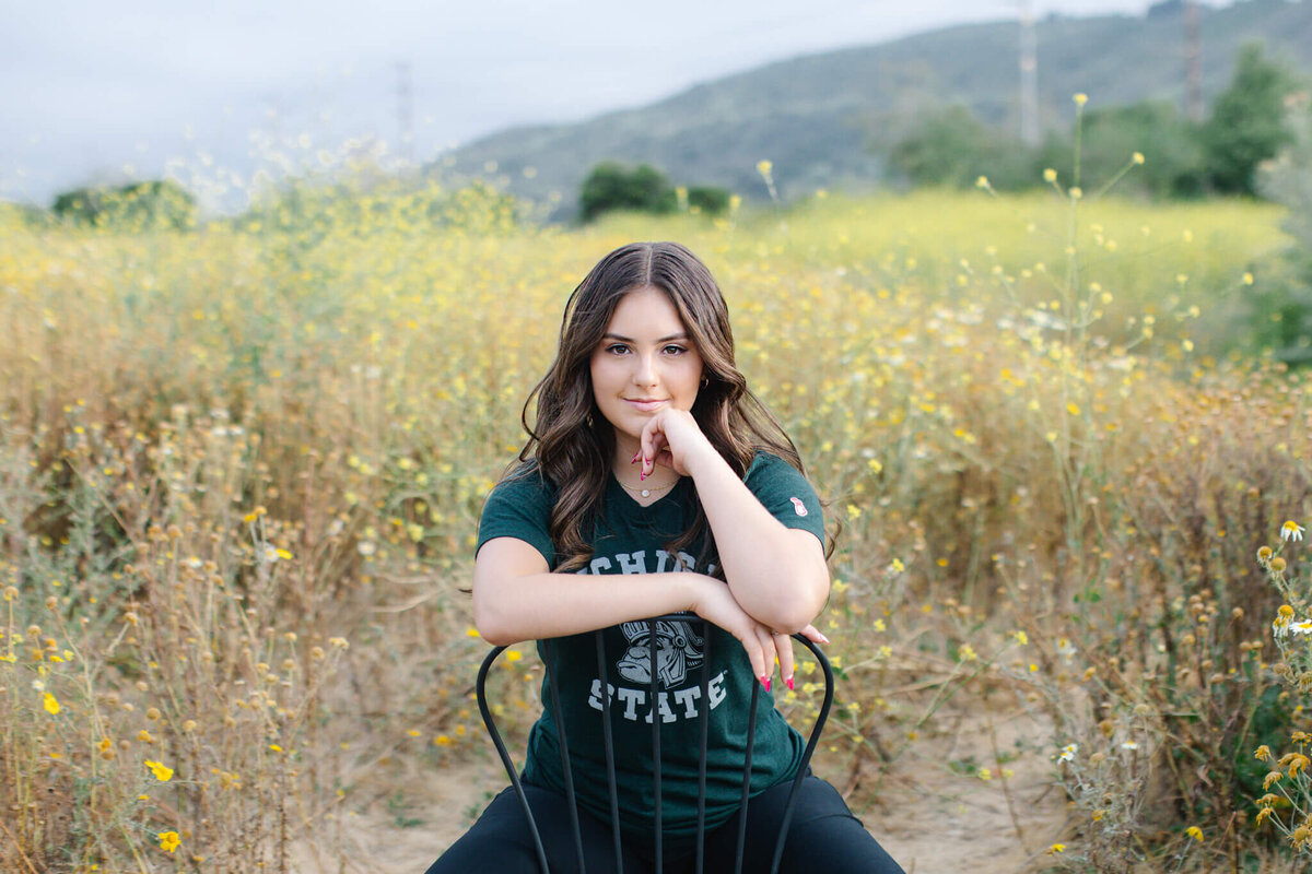 A person with long hair sits on a black chair in a field of wildflowers, wearing a dark green T-shirt with white lettering. The background features rolling hills and a cloudy sky.
