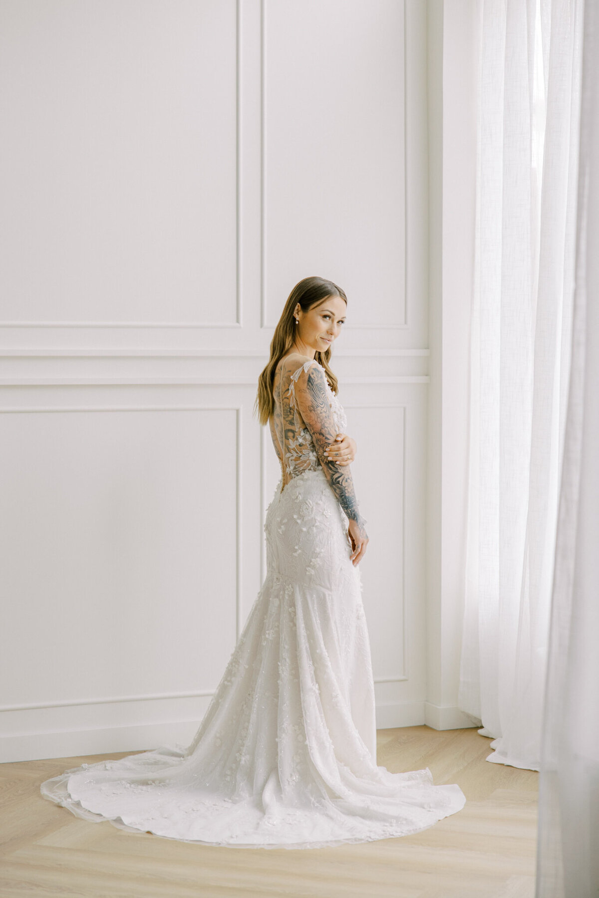 A bride in a lace wedding gown with an illusion back and floral appliqué detailing poses near a window with sheer white curtains at Studios at Vic Park in downtown Calgary. The elegant white paneled walls and natural light create a romantic and modern setting.