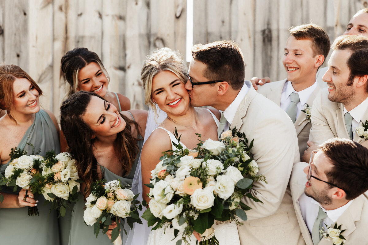 Man kisses his wife on her cheek with the wedding party.