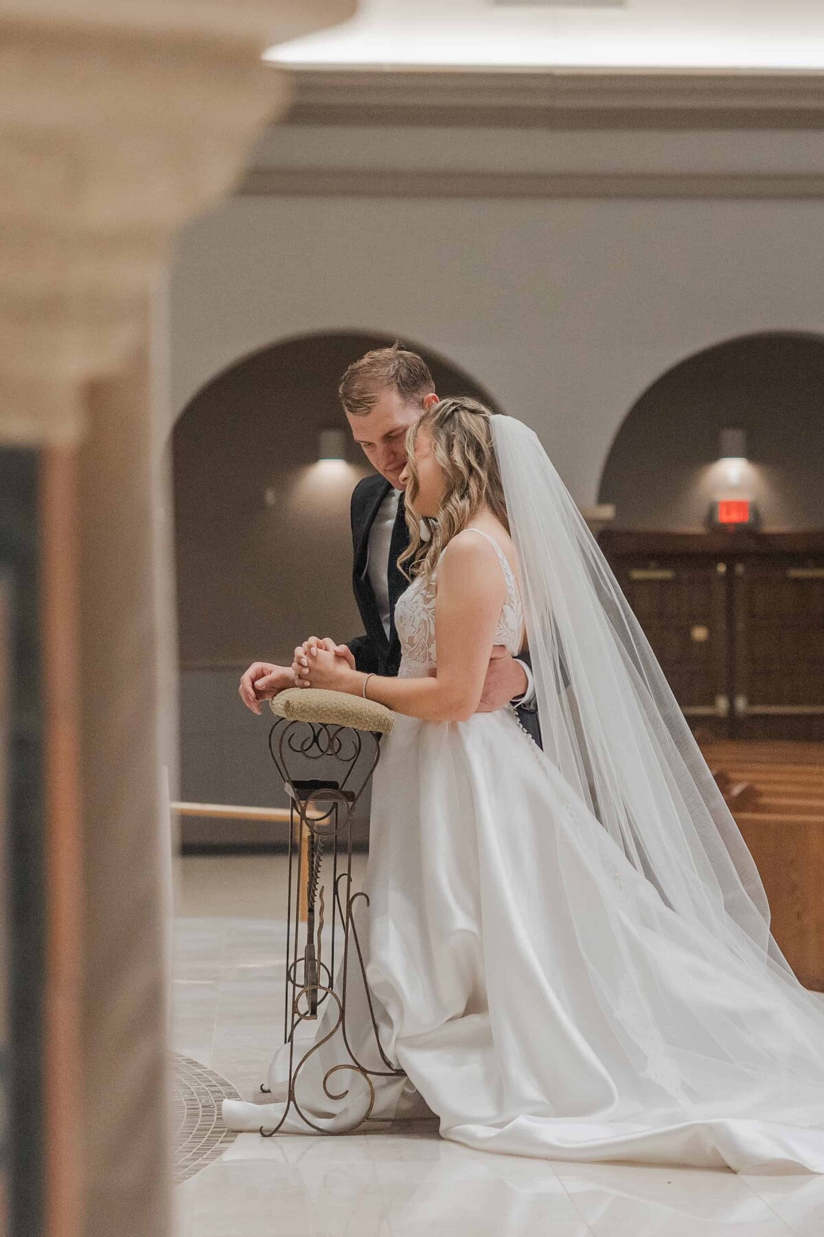 Bride and groom share an intimate prayer during their Catholic wedding ceremony at St. Joseph’s Catholic Church in Jacksonville, Florida. The bride wears a long white veil and lace bodice gown while the groom gently embraces her. Captured by Jacksonville wedding photographer Phavy Photography.