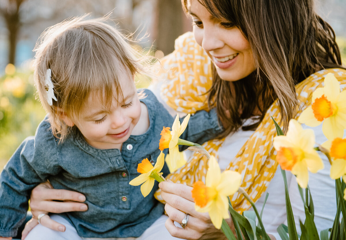 A child smiling while looking at a flower that their mother is holding.