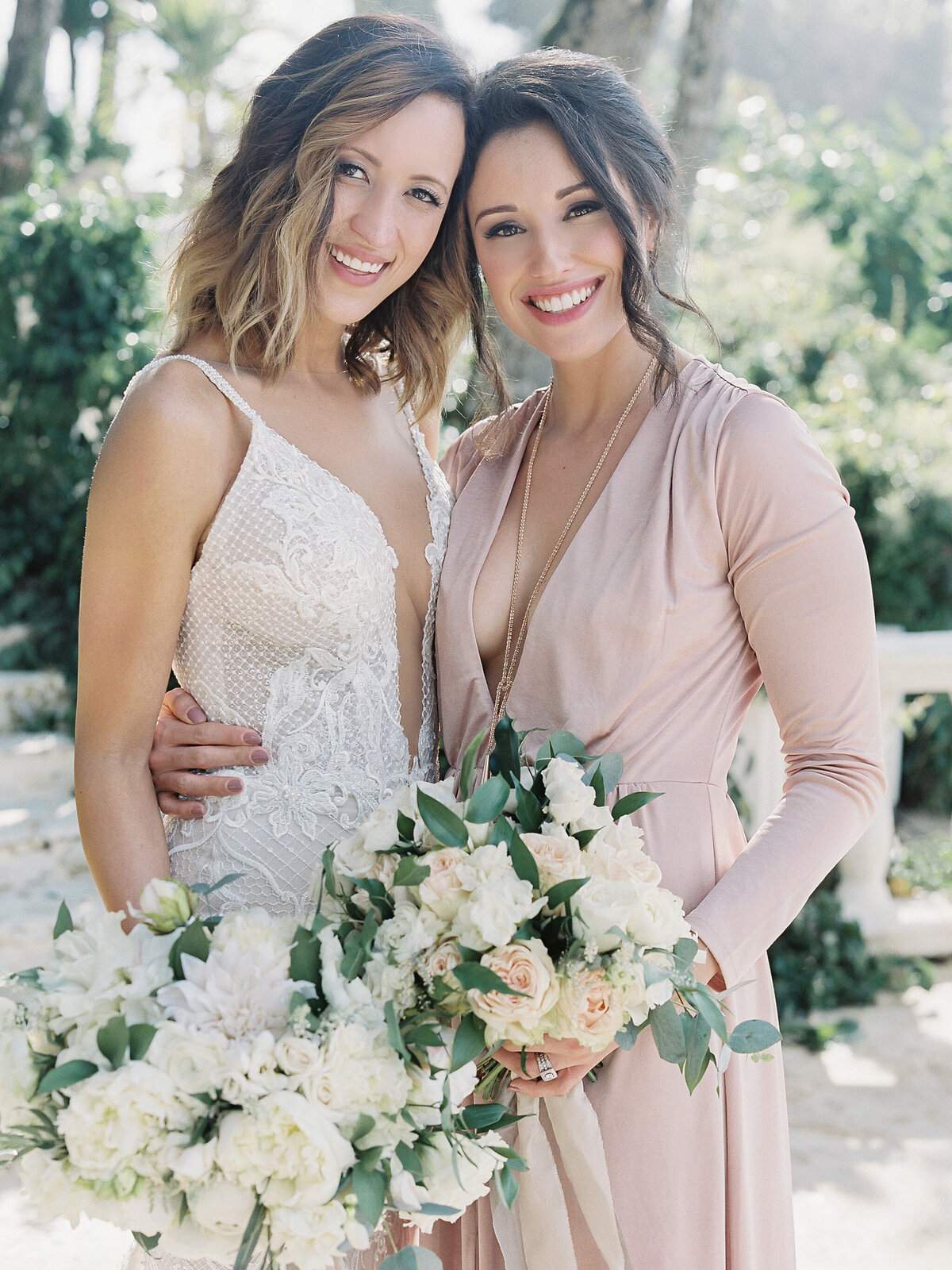 Two women are smiling and standing close together outdoors on a sunny day, holding bouquets of white and pastel flowers. One woman is wearing a white lace wedding dress, and the other is in a pale pink, long-sleeve dress. Green foliage fills the background.