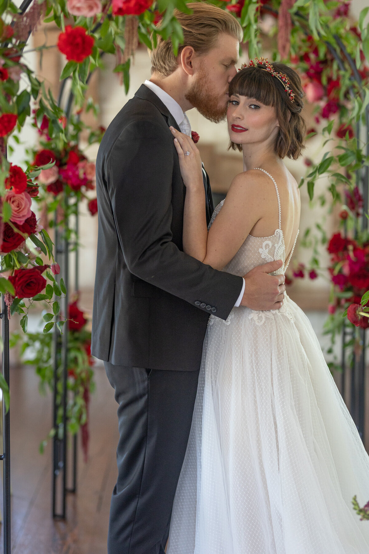 Groom kisses side of bride's head under archway of red flowers