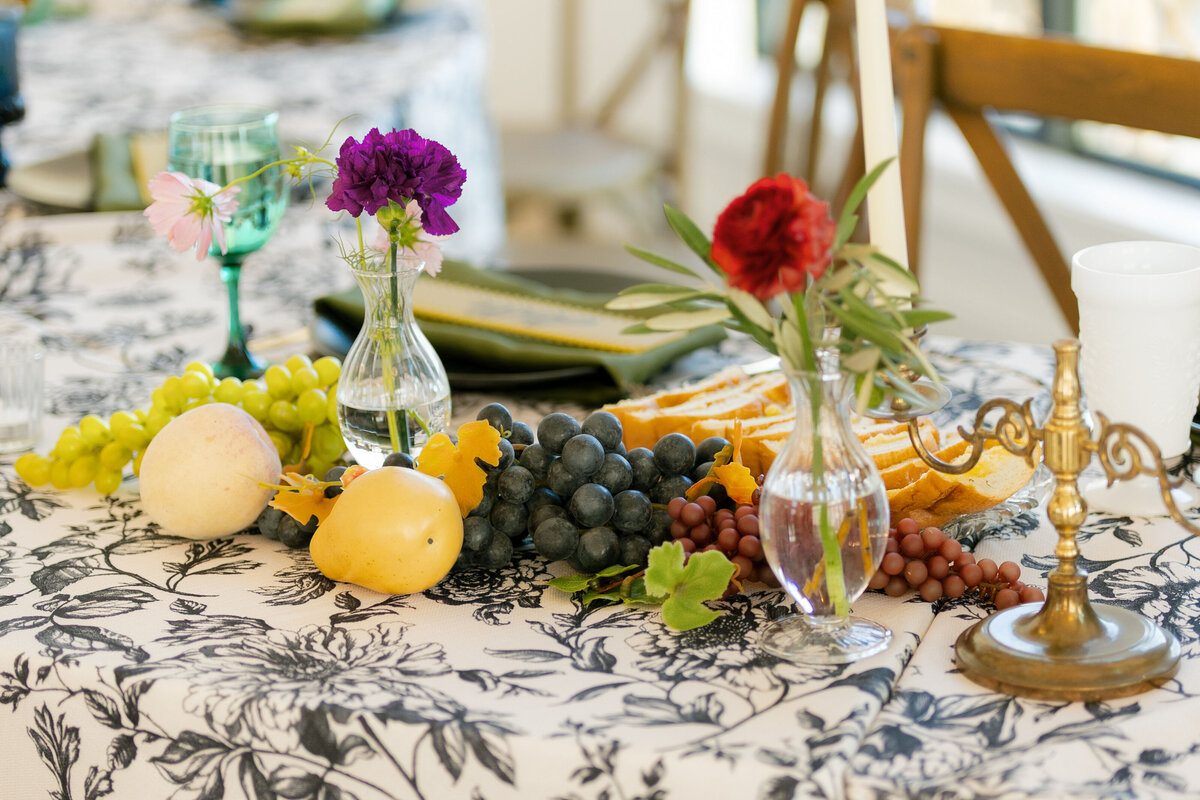 Romantic wedding centerpiece featuring colorful grapes, pears, and small florals in glass bud vases arranged across a patterned linen tablecloth for an organic, European-inspired reception design.
