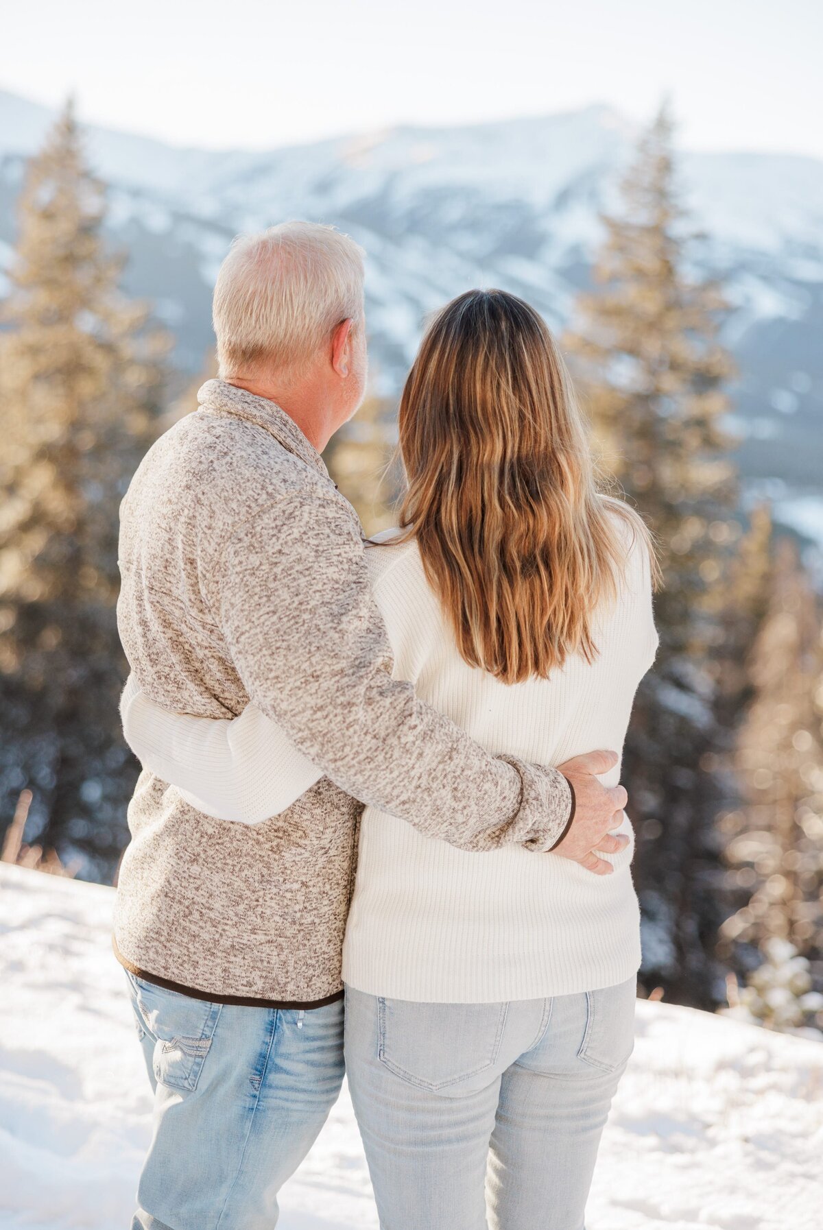 A mom and dad in sweaters hug and admire a mountain overlook in winter