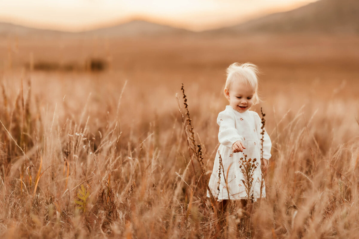 Little girl wearing white dress touches a plant in a field.