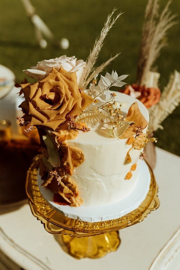 A single tier cutting cake sits on an amber glass stand. The cake is white concrete textured frosting with fresh flowers and petals in tans and white, and buttercream squiggles of terracotta frosting accents.