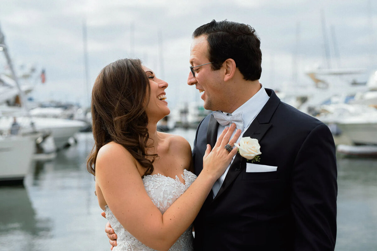 A bride and groom stand close together, smiling and laughing by a marina with boats in the background—captured beautifully by an experienced NJ wedding photographer. The bride wears a strapless white dress, and the groom dons a dark suit with a white rose boutonniere.