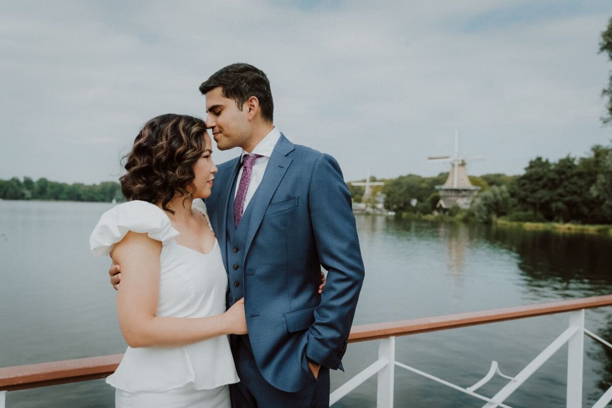  A romantic portrait of a couple standing on a wooden deck by a lake. The man, in a dark blue suit with a purple tie, lovingly kisses the forehead of the woman, who is wearing a white dress with frilly sleeves. In the background, a windmill stands on the far side of the water under a cloudy sky.