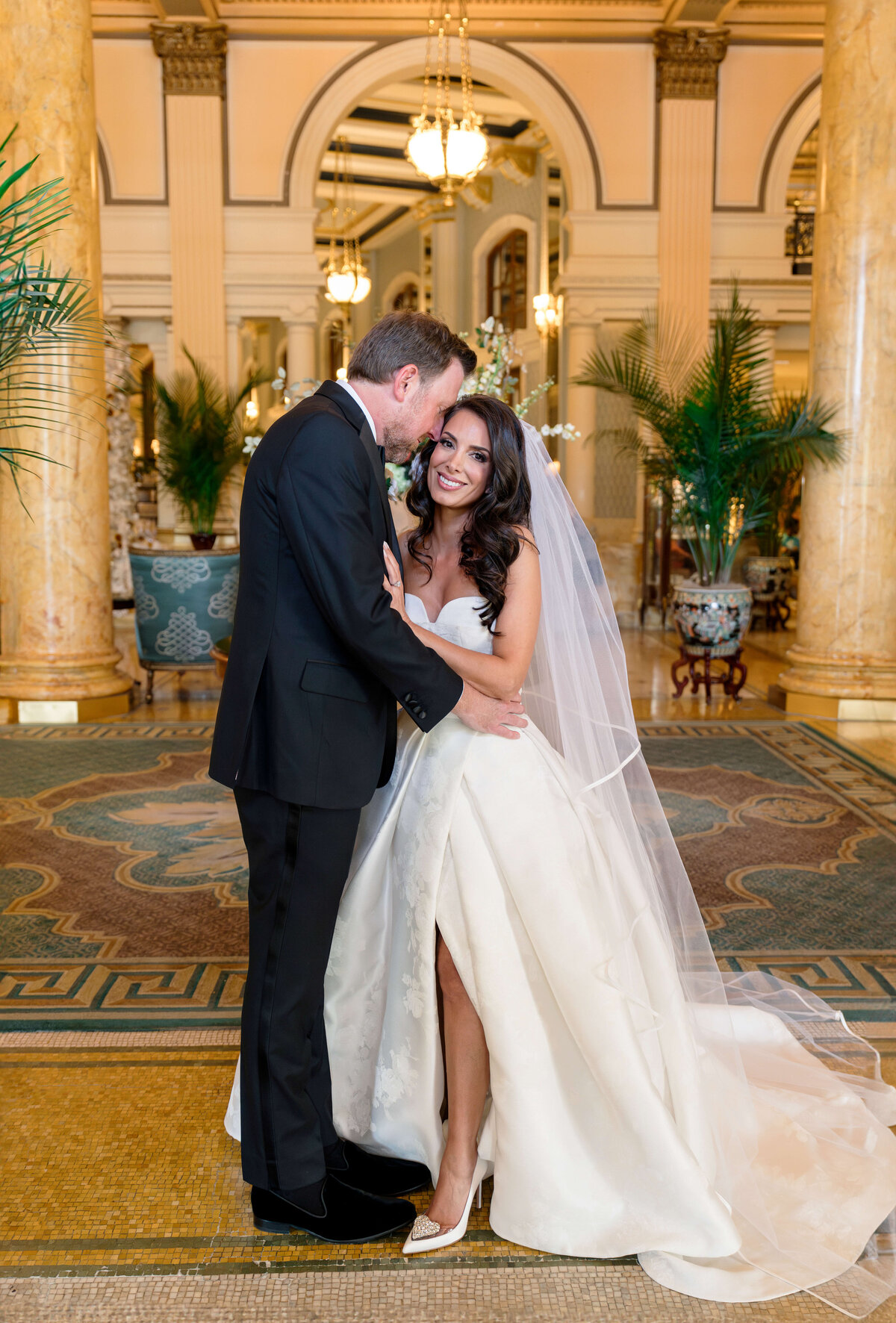 A bride and groom embrace and smile in an elegant, ornate interior with tall columns and decorative lighting. The bride wears a strapless white gown with a long veil, and the groom is in a black tuxedo.