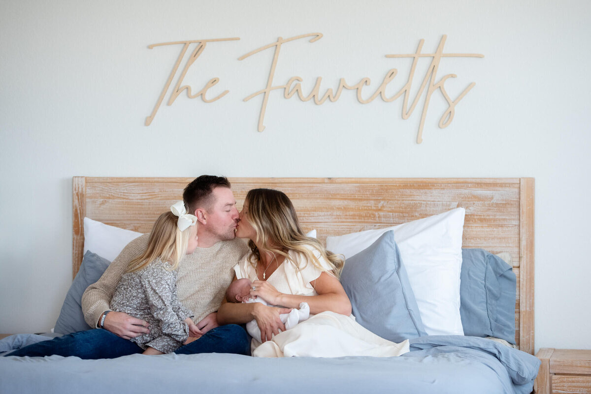 An image from a Colorado Springs Newborn Photographer of mom and dad kissing while sitting on a bed with their newborn baby and toddler daughter