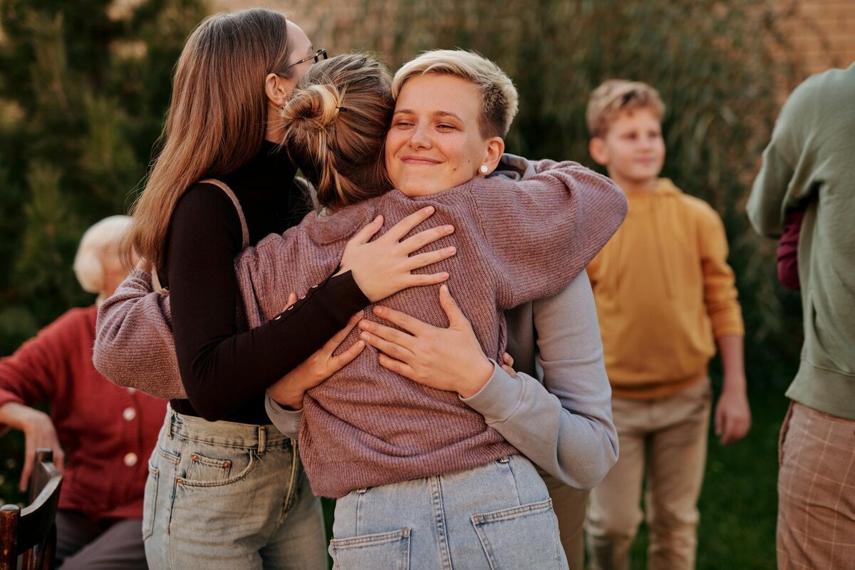 Three people are hugging and smiling outdoors, enjoying a social gathering in a garden setting. Everyone appears happy and relaxed—much like clients who connect with a therapist for teens and adults in Orange County.