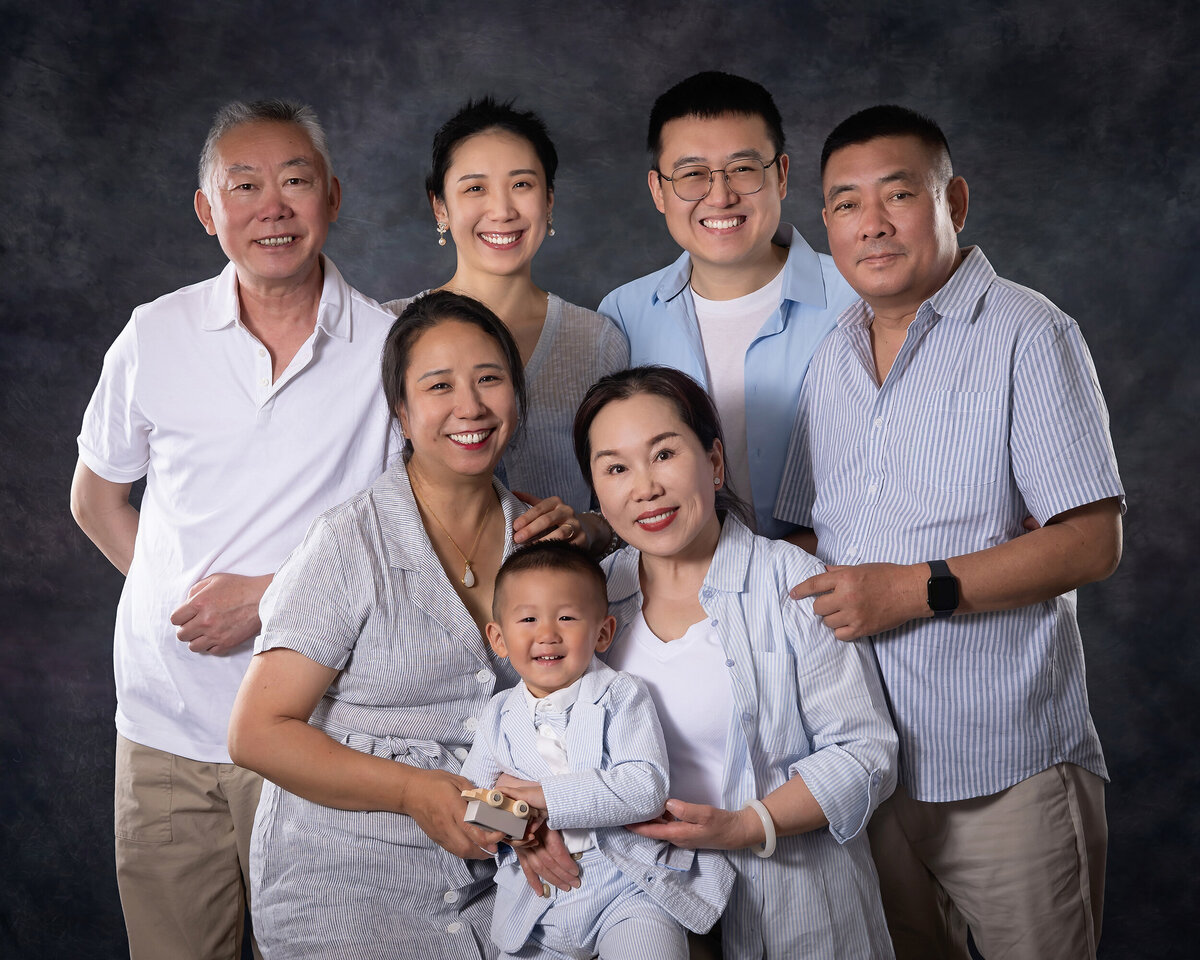 A young boy, his mom and dad, and both sets of his grandparents smile in front of a grey background. 