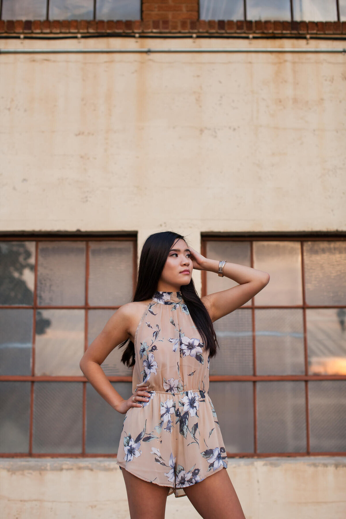 A confidently posed Asian American senior girl wearing a short dusty rose floral romper in downtown. Captured by Springfield, MO senior photographer Dynae Levingston.