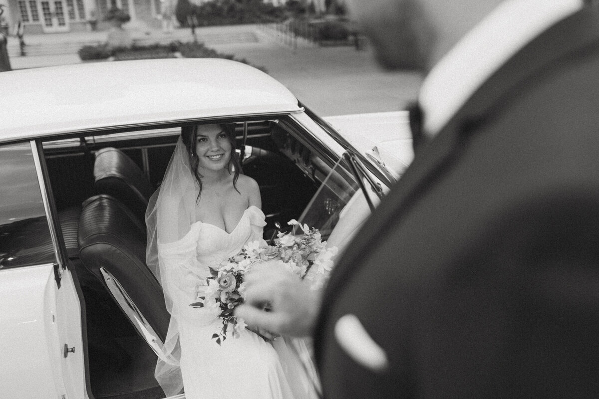A black and white photograph of a groom opening the door for his bride, beside a vintage car, evoking classic romance.