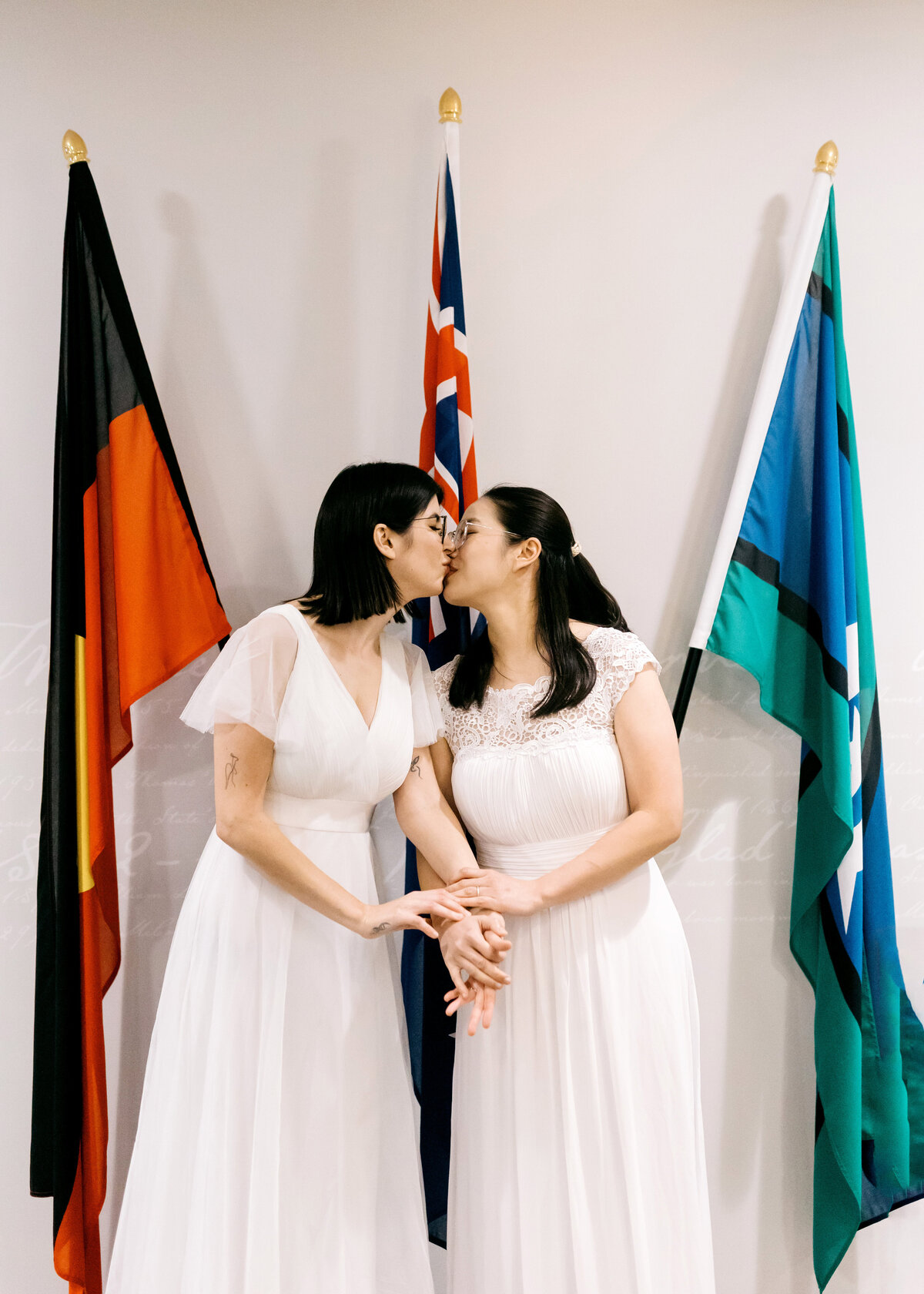 Two brides sharing a kiss after their Brisbane Registry Office wedding, standing in front of Australian and Aboriginal flags.