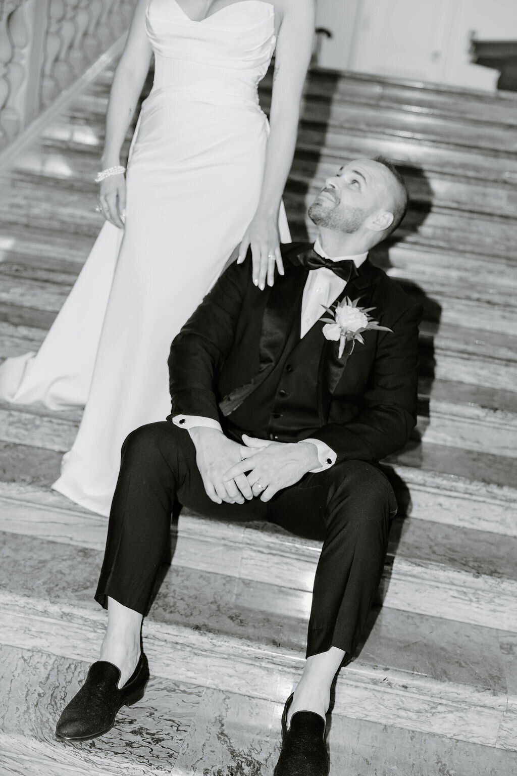 Rhode Island Wedding Photographers | A groom in a tuxedo sits on marble stairs, gazing up lovingly at a bride in a white gown. 