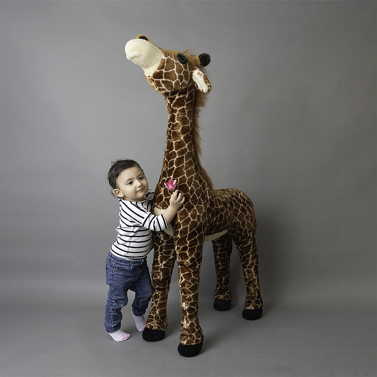 A toddler in stripe shirt and jeans hugs a large stuffed giraffe