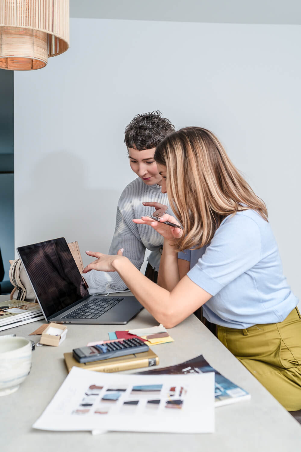 Two women reviewing design work on laptop surrounded by fabric swatches and style magazines.