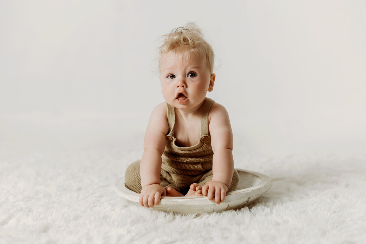 Six month old baby boy wearing a knit overalls sits in a white wooden bowl on a white rug during his Denver milestone session