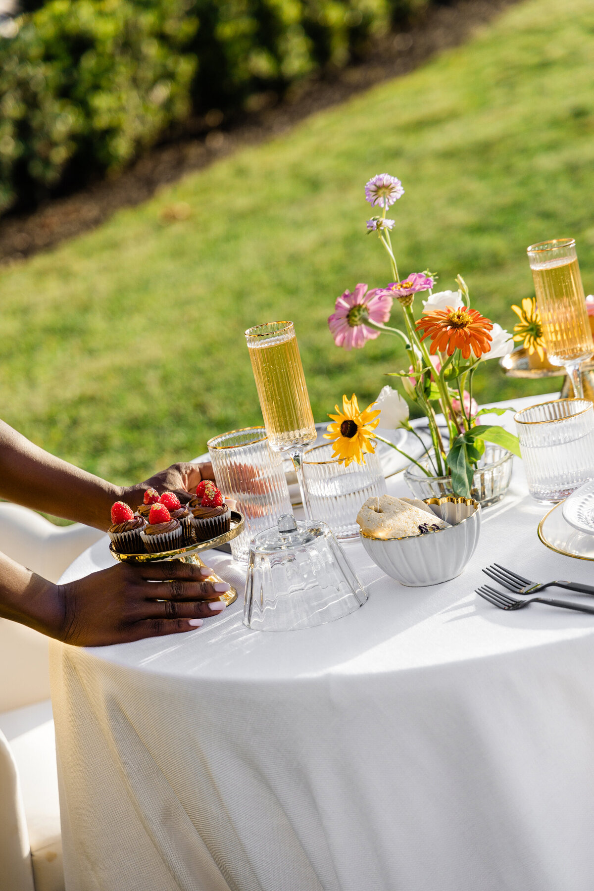 designer fixing pink orange and white flowers on white table cloth with mixed metal place setting and mini chocolate cupcakes with raspberry garnish