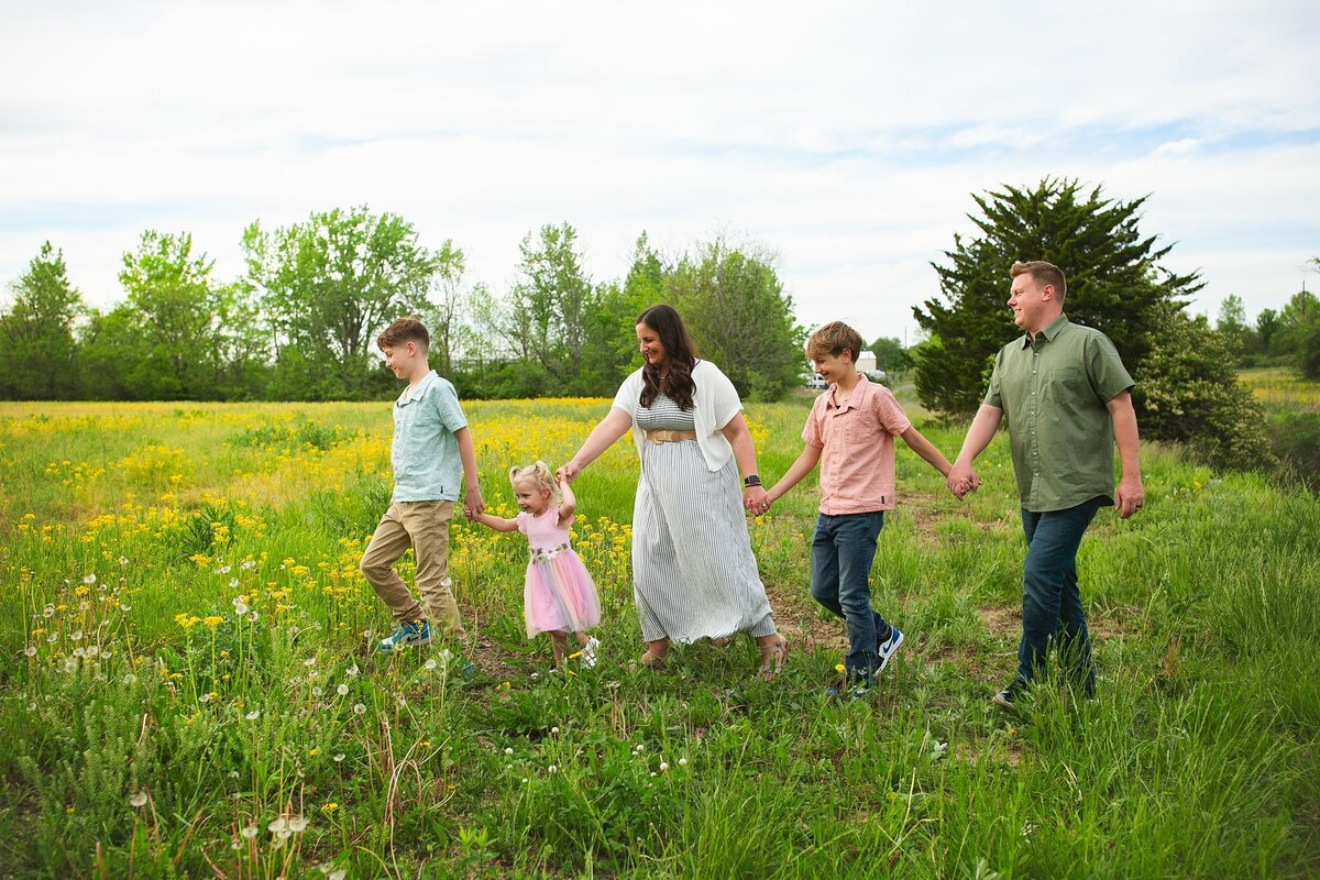 smiling-family-of-five-holding-hands-while-walking-through-a-scenic-field-of-yellow-wildflowers-in-the-center-grove-neighborhood-of-greenwood-indiana-during-a-spring-family-session