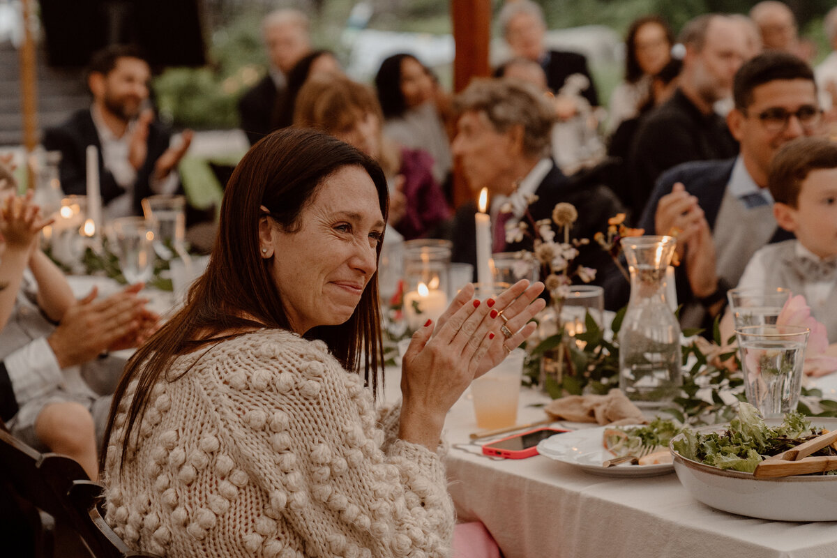 Wedding guest claps and smiles warmly during a heartfelt moment at an outdoor reception. The table is adorned with candles, floral arrangements, and dinnerware, creating an intimate and elegant atmosphere. Other guests in the background share in the joyous occasion, reflecting the celebration's warmth and community spirit.