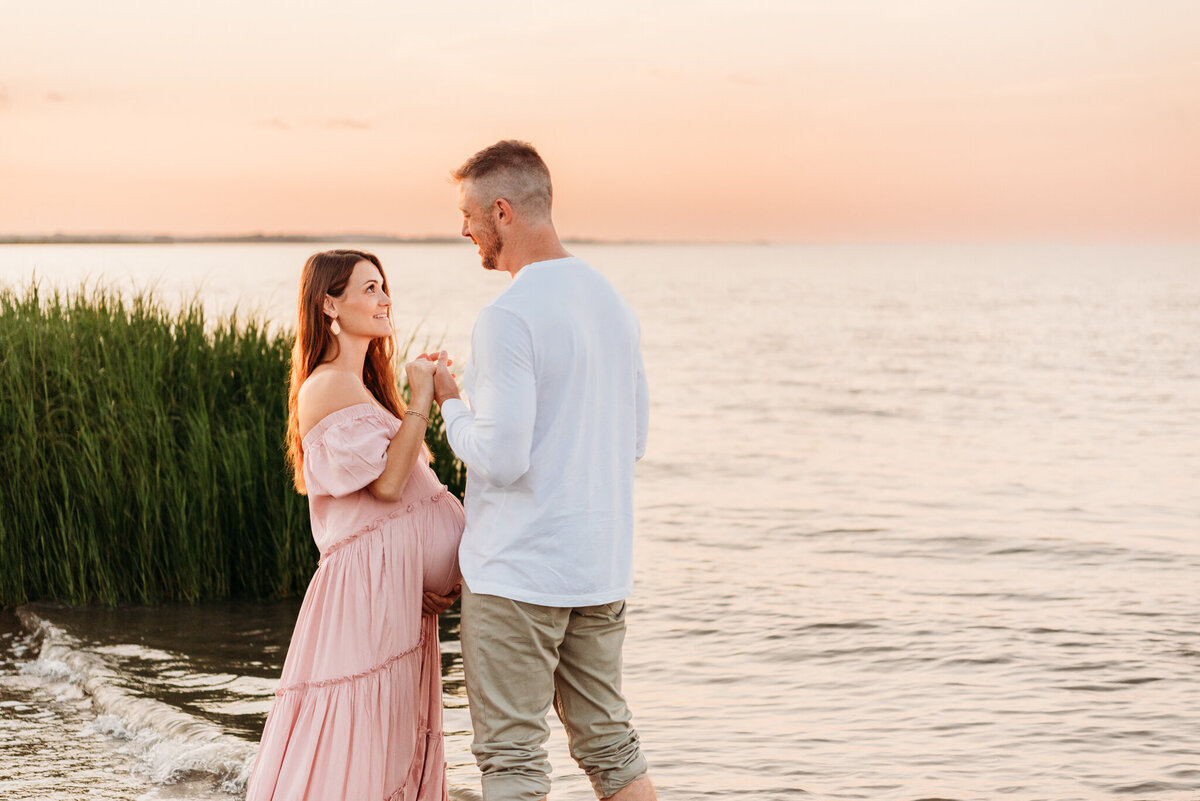pregnant woman cupping her belly from underneath, while holding her husband's hand and smiling looking into his eyes.  Photo taken during a maternity session by Cape May Photographer, Kristi