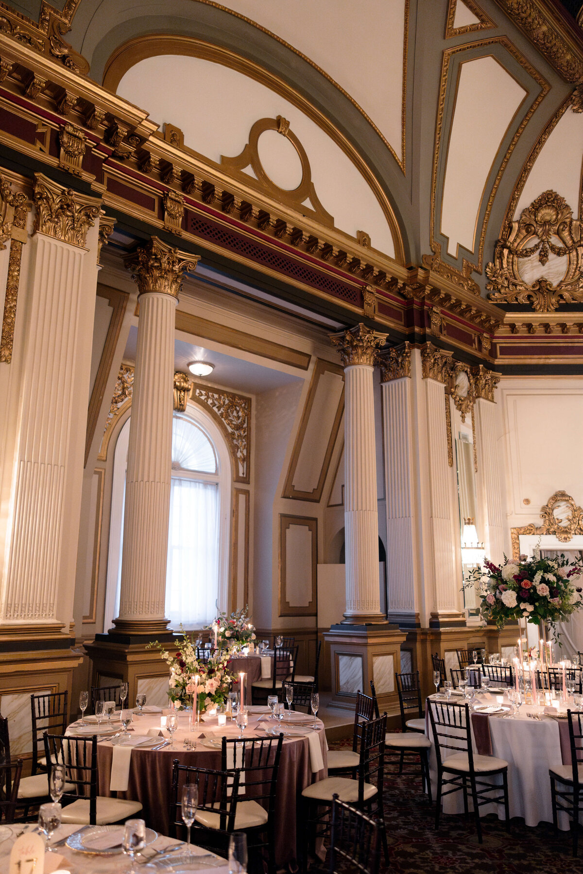 A grand ballroom with ornate gold and white classical architecture features tall columns and arched windows. Round tables are elegantly set with floral centerpieces and candles, creating a luxurious atmosphere.