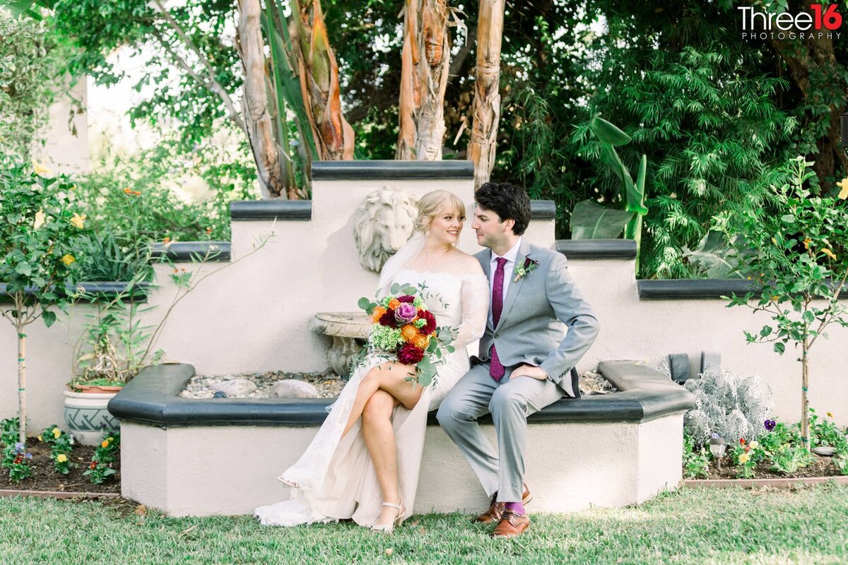 Newly married couple gaze into each other's eyes while sitting on the edge of a water fountain