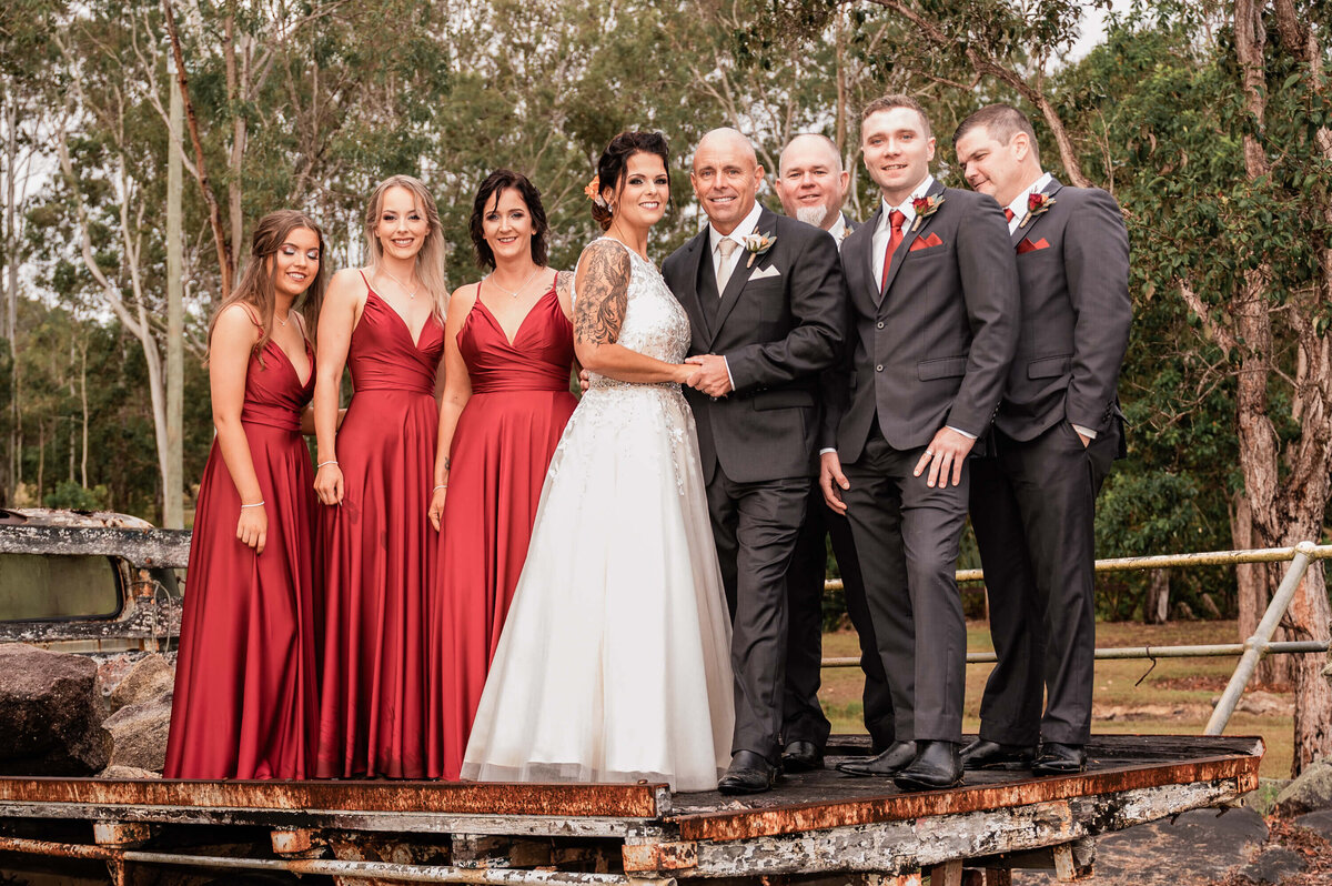 A cheerful wedding party gathers for a photo in front of a tree, celebrating their special day with smiles and laughter. - photo by Jamie Simmons