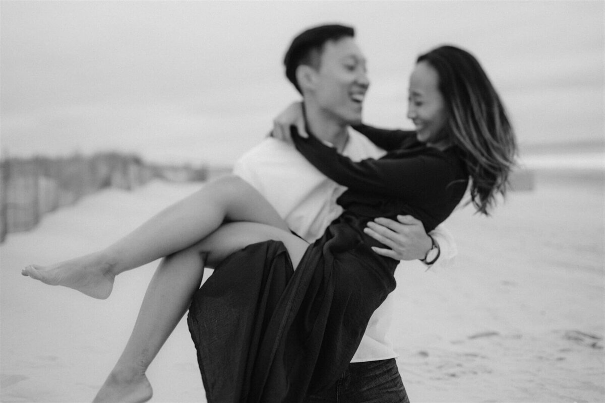 Black and white photo of a man happily carrying his sexy fiancee in Fire Island Beach, NY. Engagement Image by Jenny Fu Studio