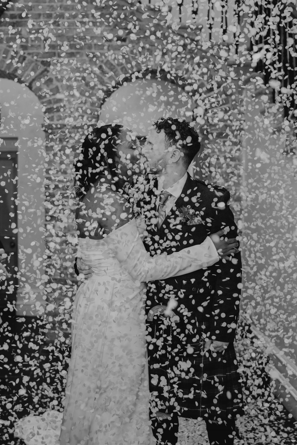 A bride and groom are photographed in the entry hall of Logie Country House as guests throw confetti from above them. The confetti falls like snow indoors and the couple smile at each other.