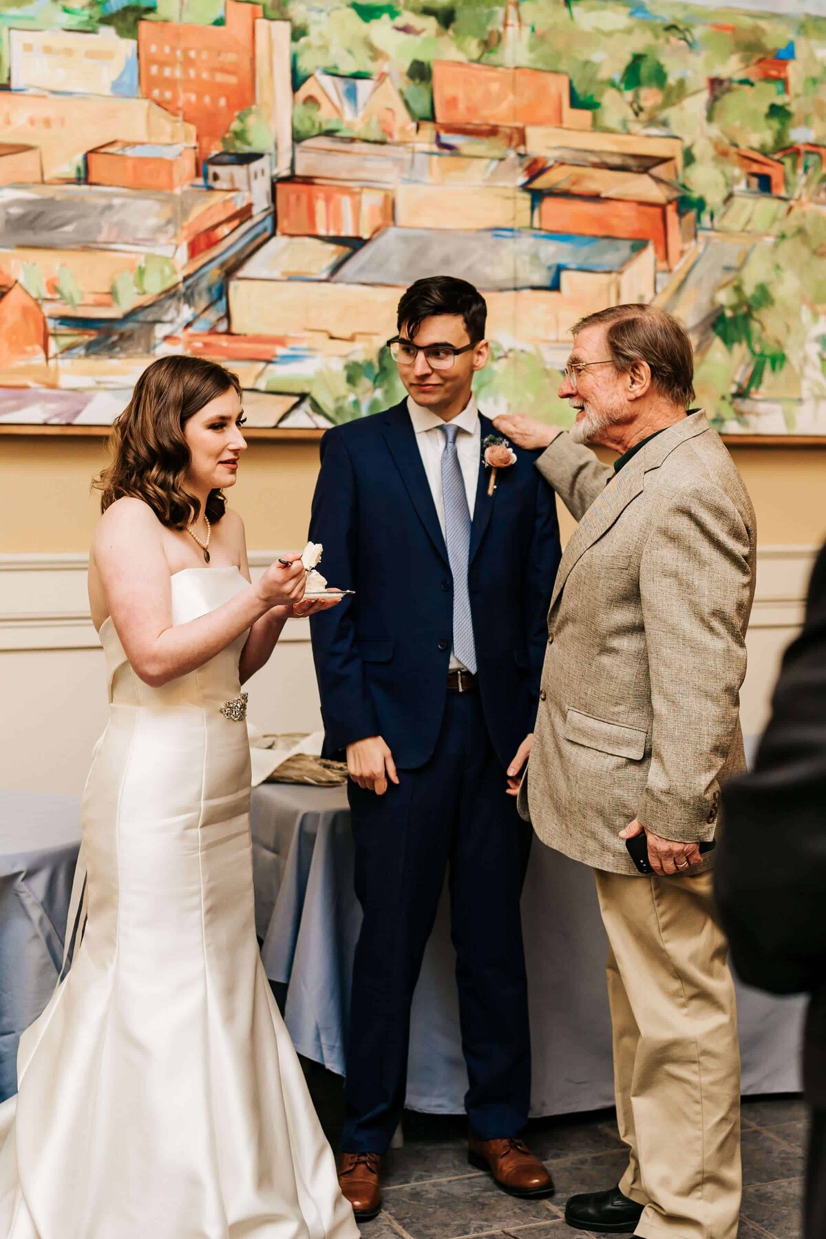 A bride in a white gown stands with a groom in a navy suit and an older man in a tan blazer. The bride holds a piece of cake, while the older man gestures toward the groom. A colorful painting hangs on the wall behind them at an indoor event.
