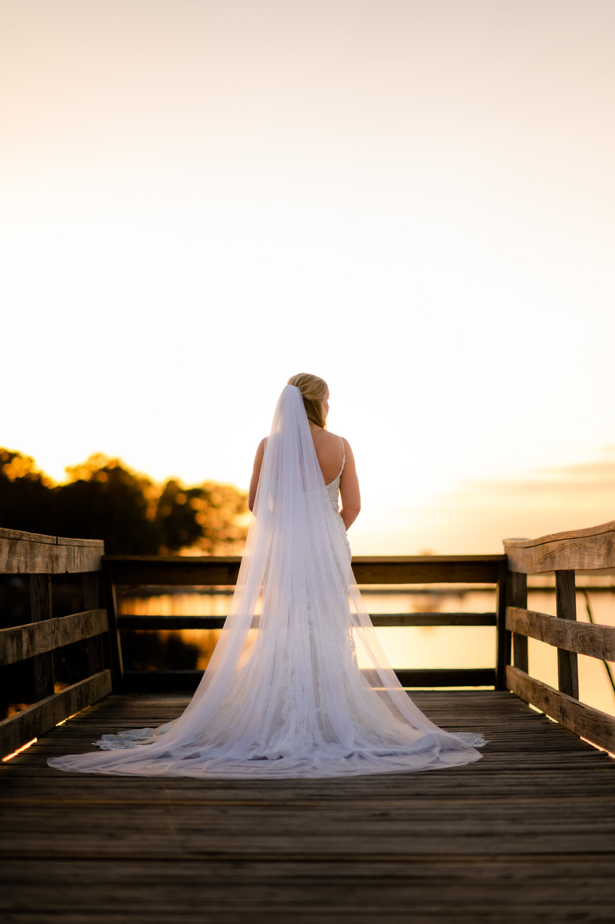 sun sets over the water at an outdoor bridal session on the lake with the brides lace train and long veil trailing behind her
