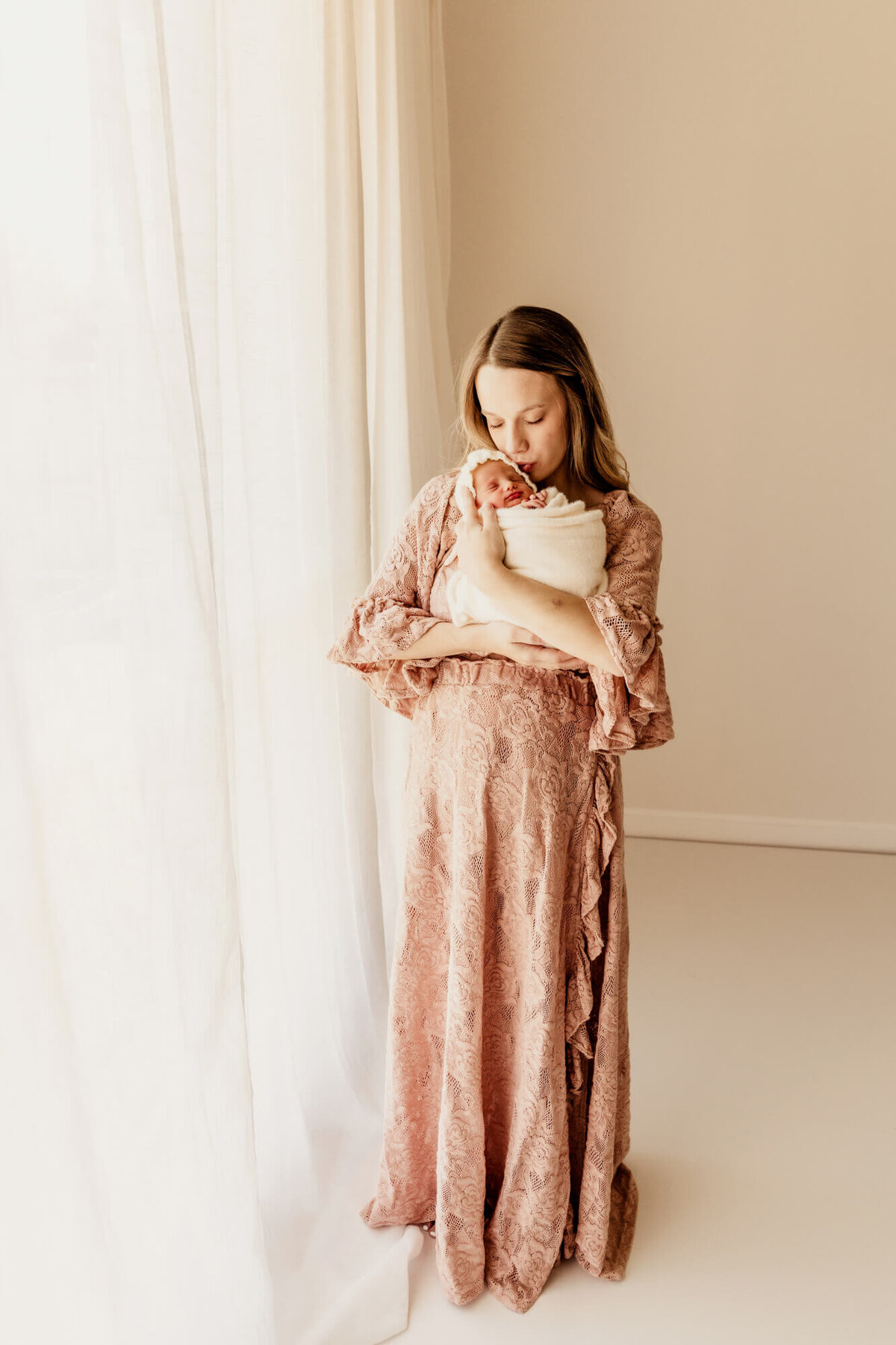 Mother wearing a hand made designer dress kisses her newborn baby girl who is wrapped in a white swaddle.