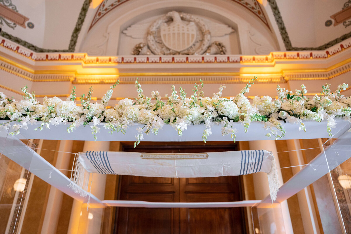 A beautifully decorated chuppah with white flowers and greenery, set under an ornate archway with intricate architectural details and warm lighting.