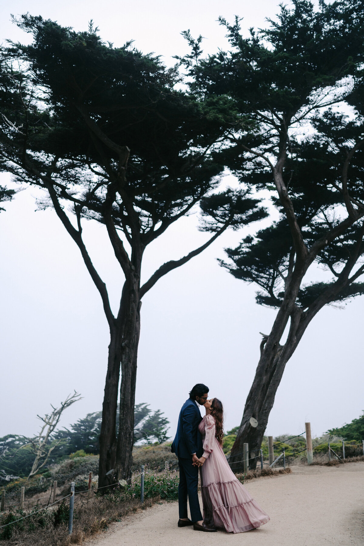 A photograph in color of Heera and Nithin during their engagement portrait session at Sutro Baths in San Francisco, California. The full length photograph shows the couple standing between two tall beach cypress trees in the midst of fog. He is wearing a blue suit and she is wearing a long sleeved, flowing, floor-length purple dress. They are facing each other, holding hands at their sides and kissing. Wedding photography taken by Stacie McChesney/Vitae Weddings.