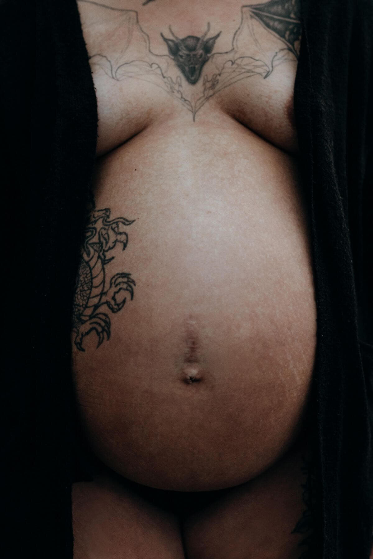 Närbild på en gravid mage med tatueringar och svart kofta under en gravidfotografering i Stockholm