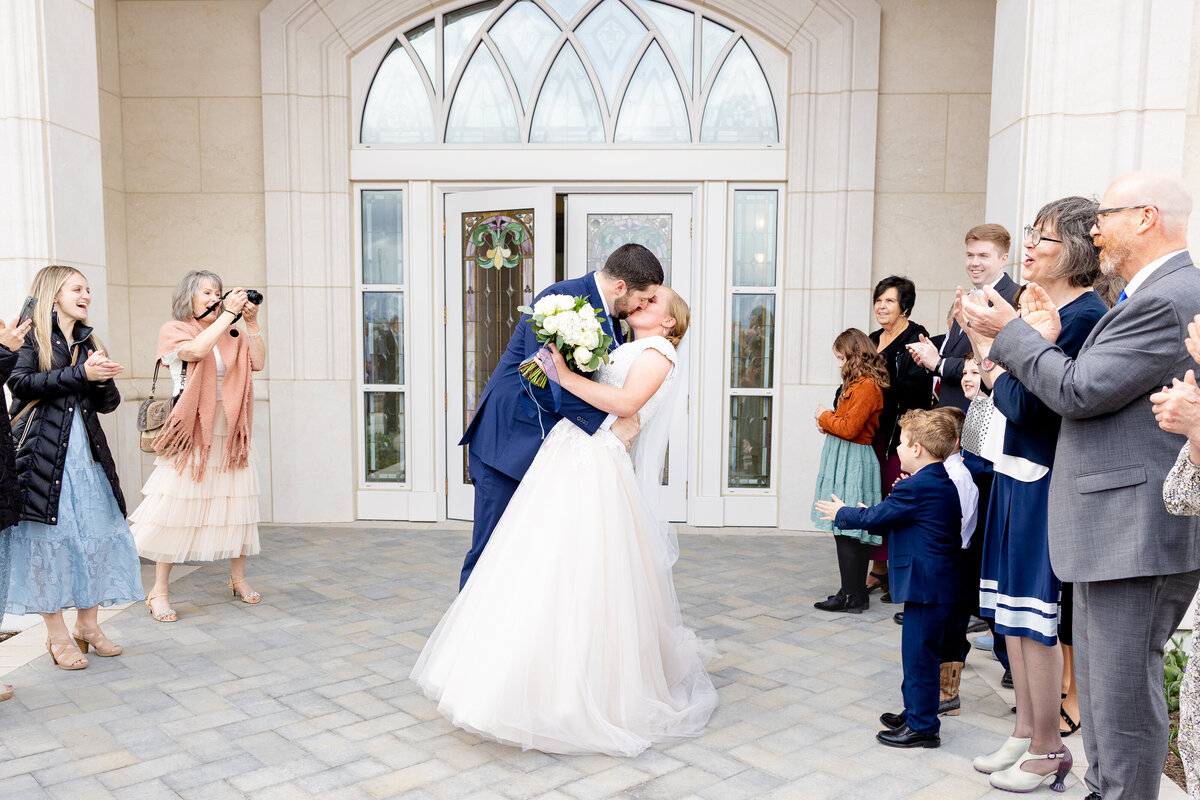 Groom dips and kisses the bride while holding a bouquet of white flowers, with loved ones clapping and smiling in the background.