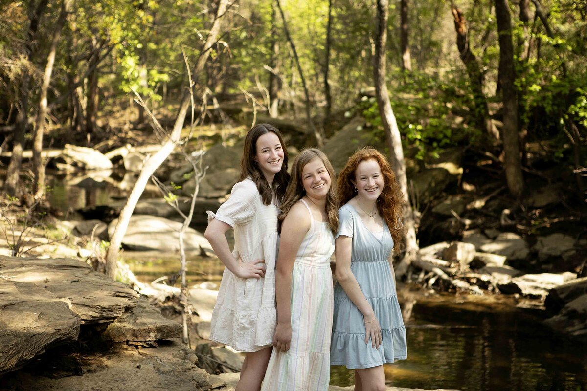 Three sisters walking hand in hand along the creek in Flower Mound, posing for a family photo surrounded by nature