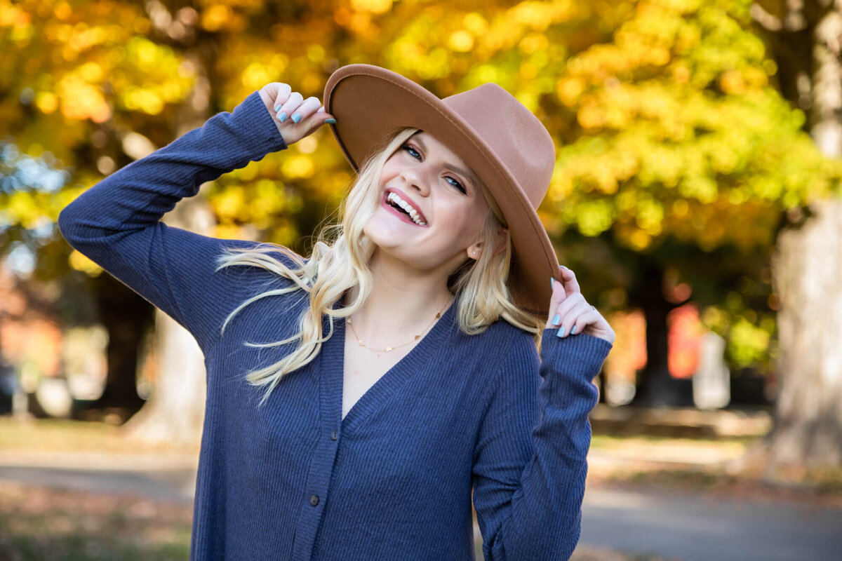 A beautiful blonde teen wearing a dusty blue top smiling and having fun posing with her wide brimmed hat. Captured by Springfield, MO senior photographer Dynae Levingston.