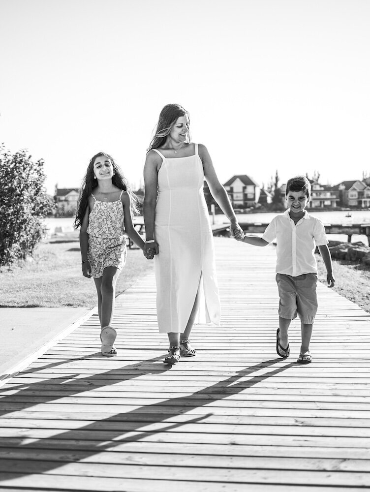 A mom in a white dress is walking near a lake holding her son and daughter's hands
