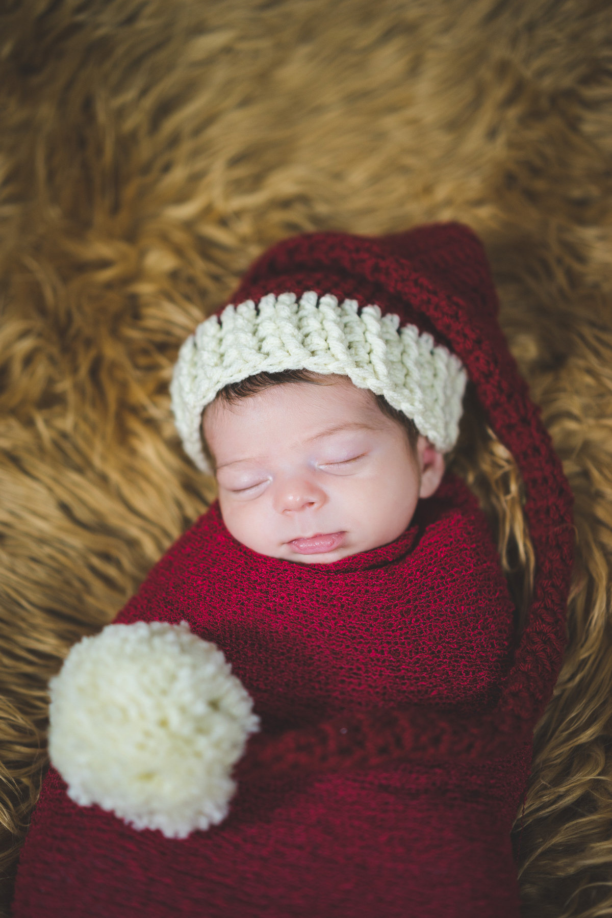 Newborn baby wrapped in red cheesecloth blanket and wearing Santa hat prop in San Antonio Photography studio.