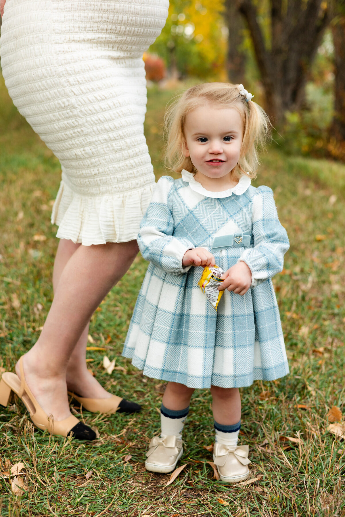A little girl stands in front of her pregnant mom and eats a snack while she looks at the camera.