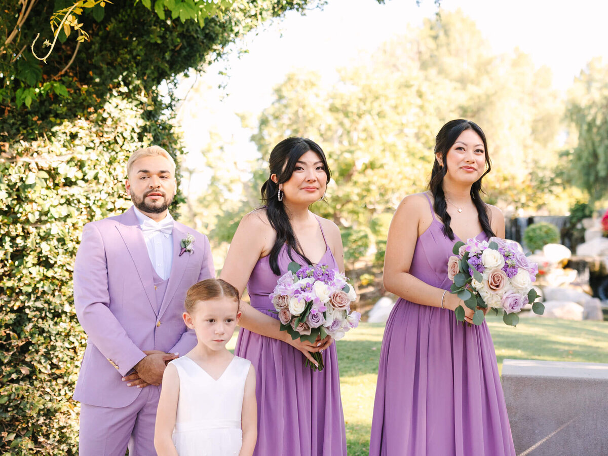 A wedding party outdoors features two bridesmaids in lavender dresses holding bouquets, a groomsman in a matching suit, and a girl in a white dress.