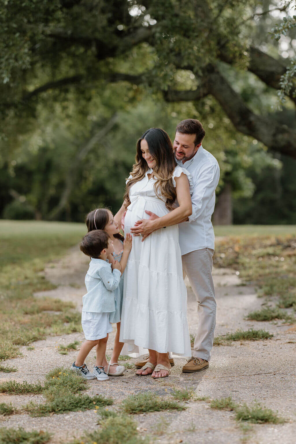 candid family picture of pregnant mother in long white dress standing with husband and 2 small children under large oak tree in Longview, TX