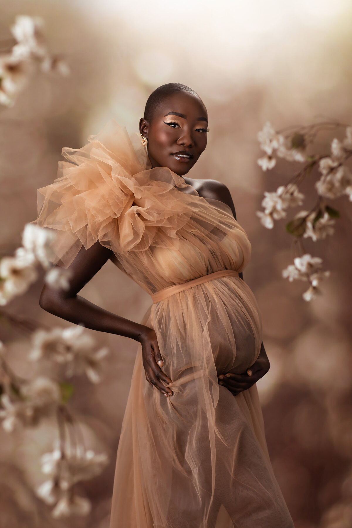 A radiant woman gracefully poses in a flowing beige gown surrounded by delicate white blossoms, embodying the elegance of DFW maternity photography.