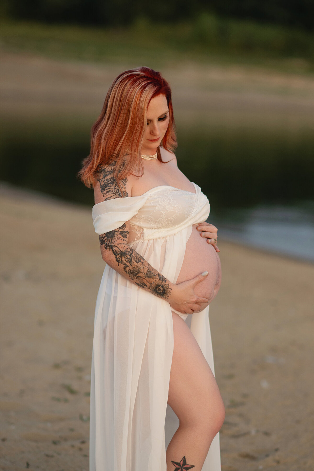 A mother to be stands on a beach in a white maternity gown at sunset gazing down at her bump in her hands